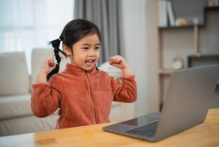 Excited Girl Celebrating Success in Front of Laptop. A happy young girl with pigtails raises her arms in triumph in front of a laptop, expressing excitement and achievement.