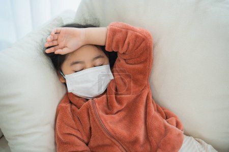 Unwohlsein asiatisches Kind ruht auf Sofa mit Maske. Ein Kind mit Gesichtsmaske ruht mit dem Arm über der Stirn auf einem Sofa, was auf Krankheit oder Unwohlsein hindeutet.