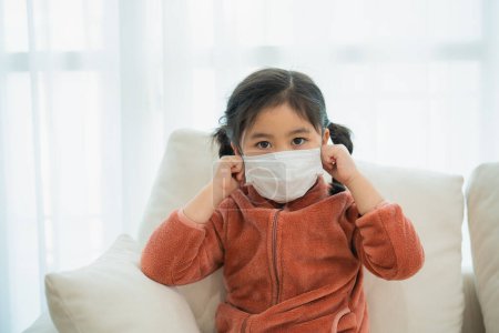 Young Asian Girl Adjusting Her Protective Face Mask. A young child in a cozy fleece adjusts her face mask while sitting on a sofa, looking directly at the camera.