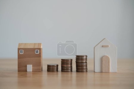 Real Estate Savings Concept with House Models and Coins. Two wooden house models beside rising stacks of coins on a clear surface, symbolizing investment growth. Finance home loan or interest concept.