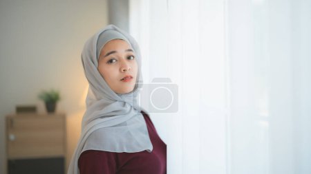 Muslim Islam woman wearing hijab or grey scarf stands in front of a window. She is smiling and looking at the camera