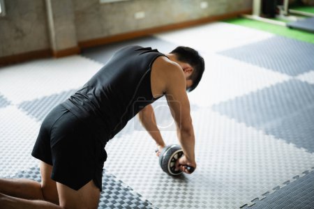 Sport man in black tank top and shorts performs an intense ab roller exercise on a patterned gym floor. His focus and determination are evident as he works out in a bright, modern gym environment.