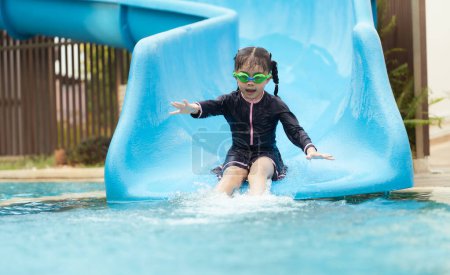 Une jeune fille glisse sur un toboggan bleu. Elle porte un maillot de bain noir et des lunettes