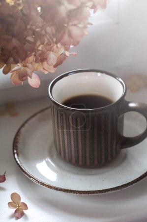 Téléchargez les photos : Une tasse de café noir sur le rebord de la fenêtre. Fleurs d'automne sèches tôt le matin. Concentration sélective à la lumière naturelle. Cadre vertical - en image libre de droit