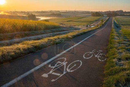 Téléchargez les photos : Route cyclable traversant une belle campagne printanière pendant la matinée brumeuse et ensoleillée - en image libre de droit
