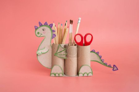 dragon shape a pencil holder made out of toilet paper and a pair of scissors, recycle concept, DIY for kids, simple activity, pink background