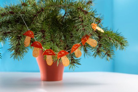 Téléchargez les photos : Un gros plan d'un arbre de Noël branches dans un pot sur une table, guirlande en écorce de mandarine déshydratée sèche en forme de champignons sur elle, art populaire, décoration artisanale d'hiver ou d'automne, bricolage - en image libre de droit