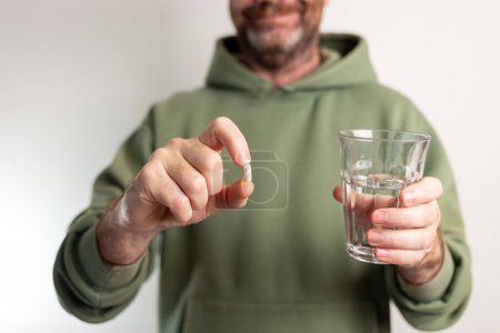 a man in a green hoodie holding a glass of water, offering the viewer a pill, soft focus
