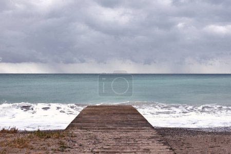 Foto de Pasarela de madera en la orilla de la playa. Nubes de tormenta, oleaje, piedras pequeñas, playa vacía, hierbas - Imagen libre de derechos