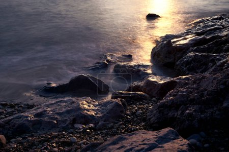 Foto de Línea de piedras de la costa al amanecer. Larga exposición, agua de efecto seda, piedras grandes y pequeñas, rayos de sol, playa vacía - Imagen libre de derechos