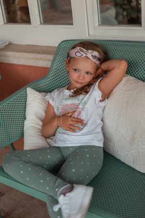 Beautiful fashionable little girl child in fashion clothes with a white T-shirt, leggings and white sneakers sits and rest on a green bench with pillows near the house