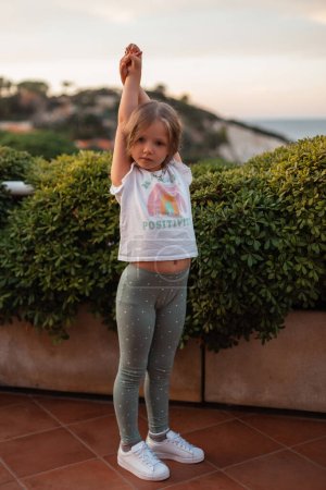 Téléchargez les photos : Belle petite fille enfant en vêtements d'été à la mode avec un T-shirt blanc, leggings verts et baskets blanches se tient près d'un buisson vert sur le bord de la mer - en image libre de droit