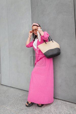 Photo for Fashionable beautiful girl plus size with a bandana in stylish summer clothes with a pink pareo and a wicker straw bag stands near a gray concrete wall and puts on sunglasses - Royalty Free Image