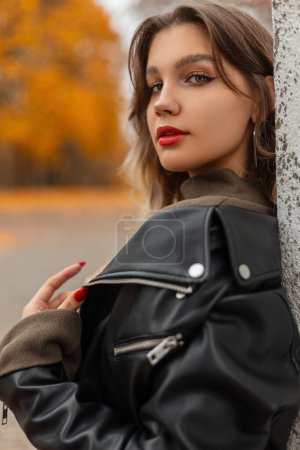 Foto de Retrato femenino otoñal de una hermosa mujer con labios rojos en una chaqueta de cuero negra de moda en un parque de otoño dorado - Imagen libre de derechos