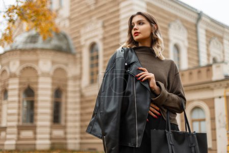 Foto de Hermosa chica joven de moda en una elegante chaqueta de cuero negro con un suéter verde con una bolsa camina en la ciudad de otoño con un edificio vintage - Imagen libre de derechos