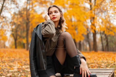 Foto de Hermosa chica de moda en ropa casual con estilo con una chaqueta de cuero, jersey y pantalones cortos se sienta en una plataforma de madera en un parque de otoño con follaje amarillo - Imagen libre de derechos