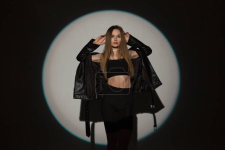 Foto de Moda hermosa chica en ropa de rock negro con estilo con una parte superior y una chaqueta de cuero posando en el estudio sobre un fondo oscuro con una luz círculo duro - Imagen libre de derechos