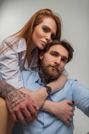 Fashionable beautiful stylish couple in love sits and hug in studio. Pretty redhead woman and hipster handsome man