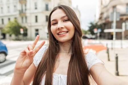 Happy beautiful young woman with a smile in a fashionable white dress walks in the city, takes a selfie photo on a smartphone and shows a peace sign
