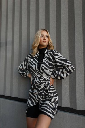 beautiful fashion blonde girl in a fashionable zebra dress with shorts stands near a white wall on the street