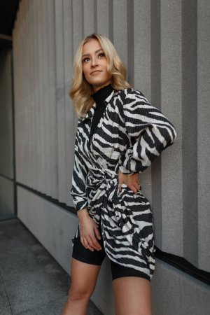 Happy fashion beautiful blondie girl in a fashionable zebra dress and shorts stands near a concrete wall in the city