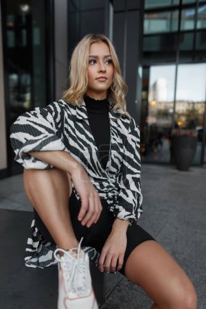 Mode schöne junge Frau Modell in modischen Kleidern mit einem Zebra-Print Kleid und weißen Turnschuhen sitzt in der Stadt in der Nähe eines Gebäudes
