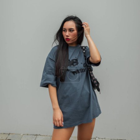 Photo for Beautiful young fashionable Asian woman model in a fashion t-shirt with shorts with a handbag stands and poses near a grey wall on the street - Royalty Free Image
