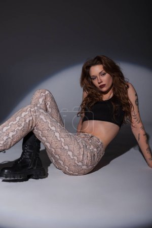 Stylish young beautiful woman in fashion clothes with top, snake print jeans and boots posing on the floor in the studio