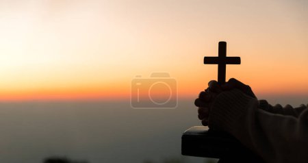 Téléchargez les photos : Silhouette de jeune femme agenouillée priant et tenant la croix chrétienne pour adorer Dieu au coucher du soleil. concept de chrétien agenouillé et priant Dieu. - en image libre de droit