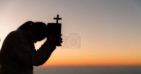 Téléchargez les photos : Silhouette de jeune femme agenouillée priant et tenant la croix chrétienne pour adorer Dieu au coucher du soleil. concept de chrétien agenouillé et priant Dieu. - en image libre de droit