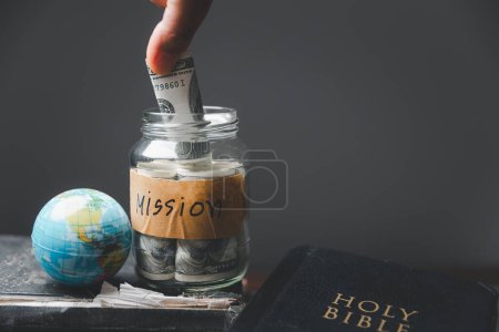 Gläser voller Geld und Globus mit der Heiligen Bibel für die Mission, christliche Mission. Hand hält Dollar mit Bibel auf Holztisch, christlicher Hintergrund für großen Auftrag oder Earth Day Konzept.