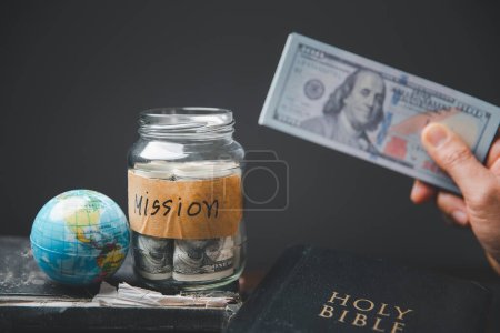 Photo for Saving jars full of money and globe with Holy Bible for mission, Mission christian idea. Hand holding dollar with bible on wooden table, Christian background for great commission or earth day concept. - Royalty Free Image