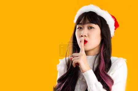 Photo for Young asian pretty woman in white blouse wear santa hat finger on mouth secrete sign yellow background. - Royalty Free Image