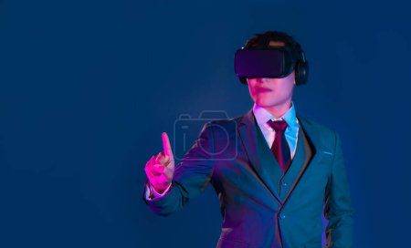 Téléchargez les photos : Asain homme en costume d'affaires portant vr lunettes de réalité virtuelle casque regarder jouer et toucher le doigt sur fond de couleur bleu foncé. Concept Metaverse - en image libre de droit