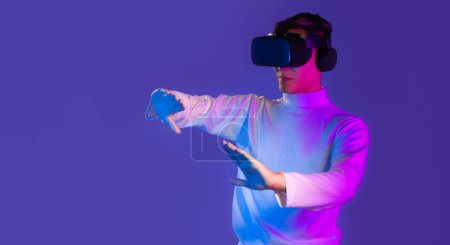 Téléchargez les photos : Homme asiatique en blanc manches longues t-shirt porter vr lunettes casque regarder jouer jeu touchant en ligne fond violet. Métaverse concis - en image libre de droit