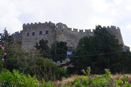 Die griechische Flagge weht vor dem Hintergrund der Festung auf der Akropolis von Lindos, die von den Rittern des Ordens des Heiligen Johannes von Jerusalem auf der antiken griechischen Akropolis aus dem 14. Jahrhundert erbaut wurde. Lindos, Insel Rhodos, Dodekanes, Griechenland