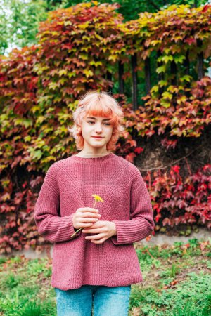 Téléchargez les photos : Jeune fille rousse debout contre feuillage d'automne coloré et tenant fleur jaune. - en image libre de droit