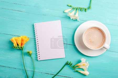 Notizblock und Kaffeetasse leer auf einem türkisfarbenen Holztisch mit Freesiablüten. Draufsicht, flache Lage, Attrappe.