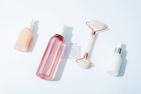 Botellas cosméticas rosadas y rodillo facial sobre fondo blanco con sombras nítidas. Cosmética natural, concepto de cuidado de la piel. Vista superior, plano.