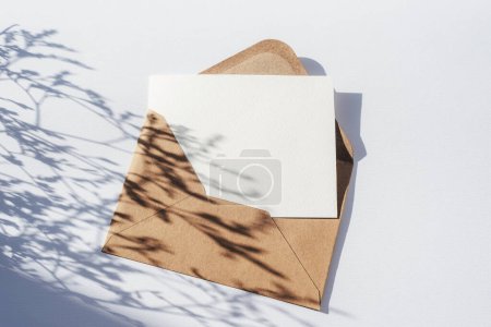 Envelope with blank card and flowers shadow on light blue background. Top view, flat lay, mockup.