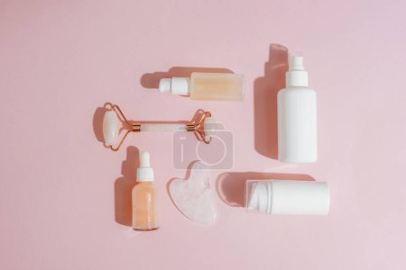 Cosmetic bottles and face roller, gua sha stone on pink background in sunlight. Skin care, spa and wellness concept. Top view, flat lay.
