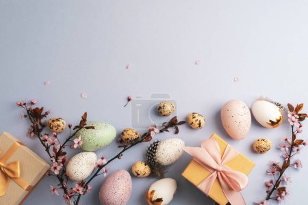 Téléchargez les photos : Pose plate de Pâques avec des cadeaux enveloppés, des œufs peints, des fleurs de cerisier et des plumes sur fond bleu clair. Vue du dessus, espace de copie. - en image libre de droit