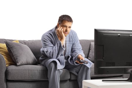 Photo for Bored young man in a bathrobe watching tv and sitting on a sofa isolated on white background - Royalty Free Image