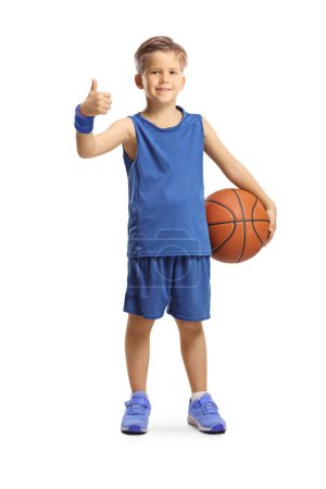 Retrato completo de un niño en un jersey azul sosteniendo una pelota de baloncesto y mostrando los pulgares hacia arriba aislados sobre fondo blanco