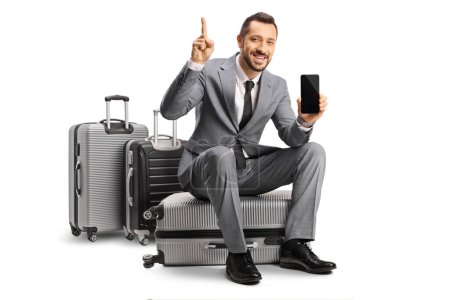 Photo for Bussinessman sitting on a suitcase pointing up and showing a smartphone isolated on white background - Royalty Free Image