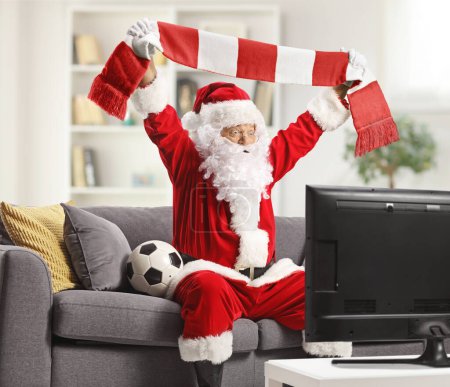 Foto de Emocionado Santa Claus viendo fútbol en la televisión y animando con una bufanda en casa - Imagen libre de derechos