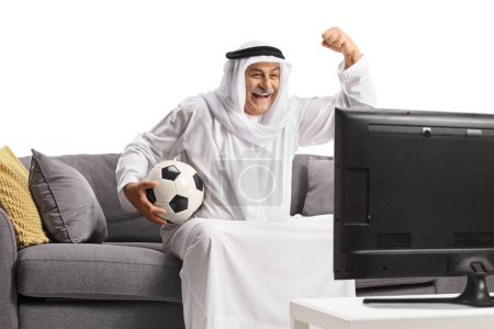 Foto de Hombre árabe maduro en una bata sosteniendo un balón de fútbol y animando sentado en un sofá frente a la televisión aislado sobre fondo blanco - Imagen libre de derechos