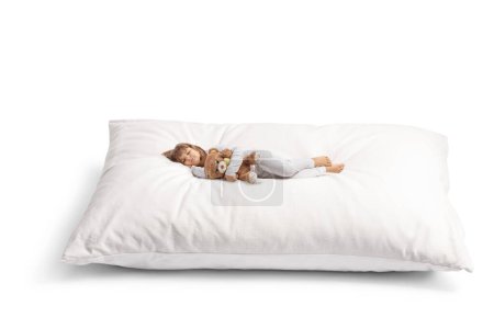 Téléchargez les photos : Petite fille en pyjama embrassant un ours en peluche et dormant sur un grand oreiller doux isolé sur fond blanc - en image libre de droit