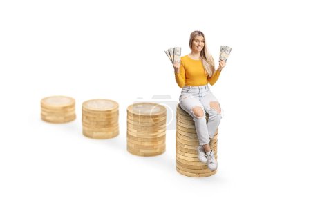 Photo for Excited young female sitting on a pile of coins and holding paper banknotes isolated on white background - Royalty Free Image