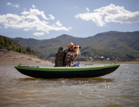 Téléchargez les photos : Homme avec un bakcpack utilisant des jumelles sur un canot dans un lac de montagne - en image libre de droit